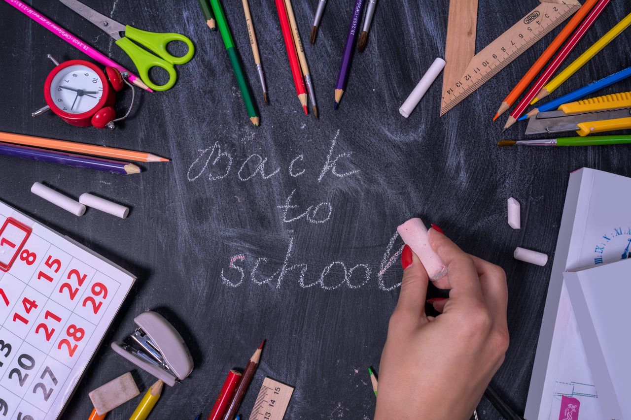 Hand writing 'back to school' on chalkboard surrounded by school supplies.
